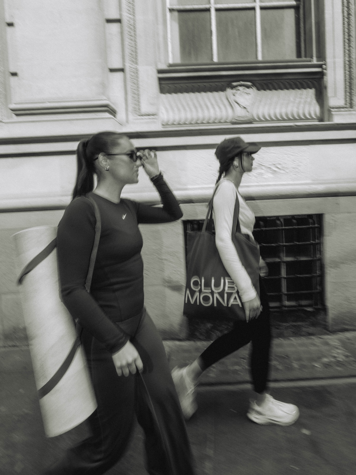 Two women walking beside a Club Mona poster in black and white