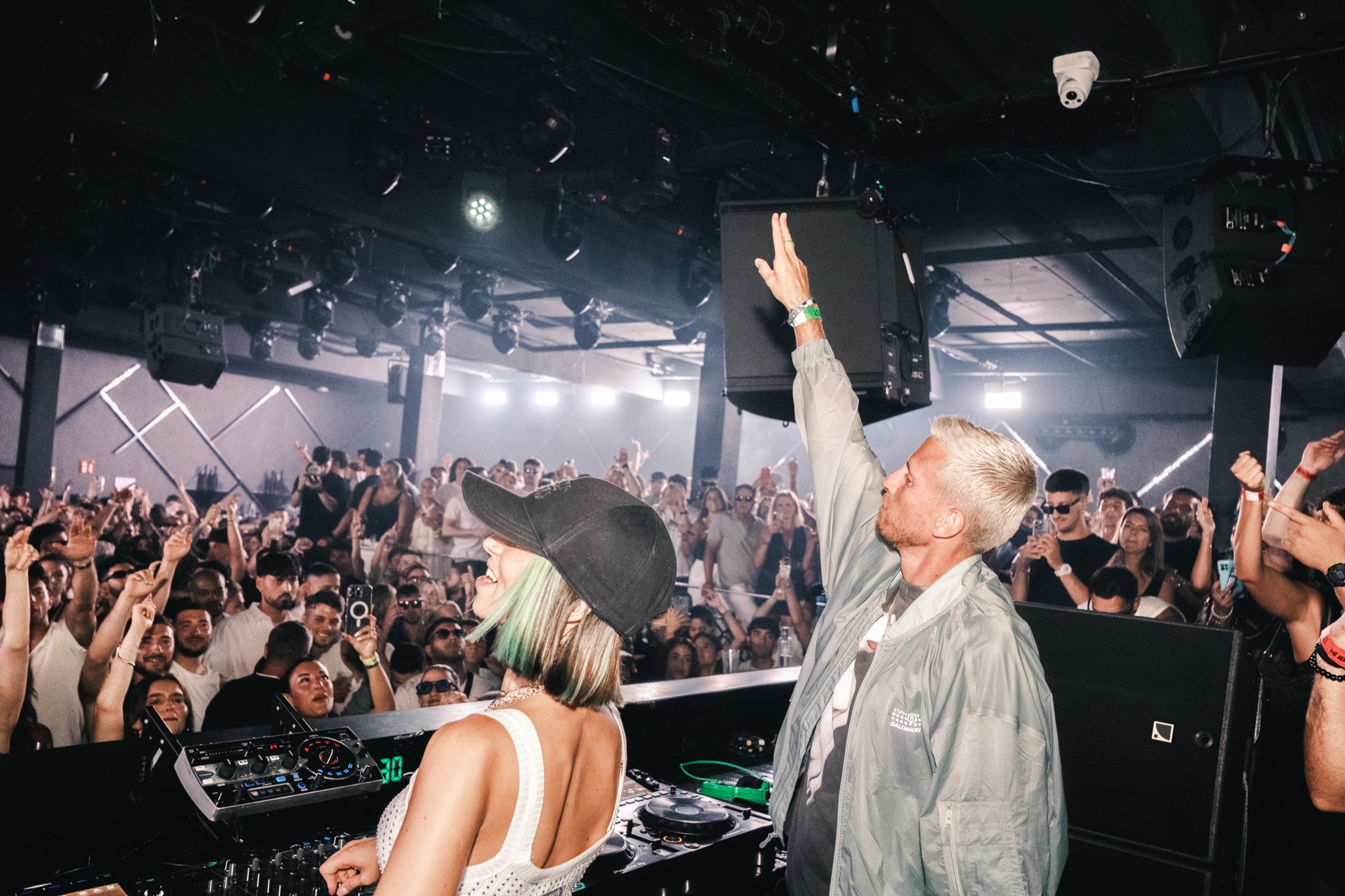 Eli Brown raising his arm in the DJ booth with the crowd behind him.
